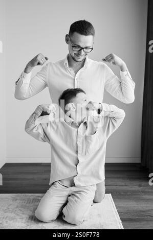 Junger Vater in Brille, der mit seinem neugierigen Sohn spielt. Lustiger Junge, der seinem Vater Muskeln zeigt. Familie genießt Aktivitäten zu Hause. Schwarzweißbild. Stockfoto