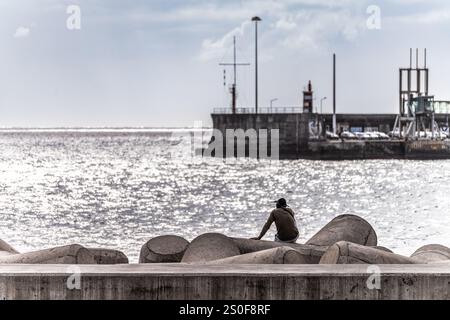 Eine ruhige Szene einer Person, die am Ufer sitzt und den Ozean mit einem Hafen im Hintergrund betrachtet. Das Bild vermittelt Einsamkeit, Entspannung, A Stockfoto