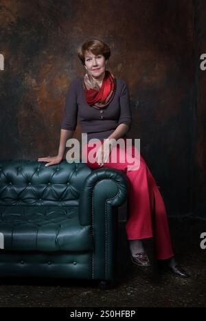 Eine Frau mittleren Alters in roter Hose saß auf der Rückseite des Sofas auf braunem Hintergrund Stockfoto
