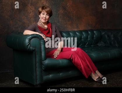 Frau mittleren Alters in roter Hose saß auf der Rückseite des Sofas auf braunem Hintergrund Stockfoto