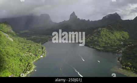 Boote, die die Bucht von Opunohu, grüne Regenwälder und vulkanische Gipfel in dramatischen Regenwolken segeln, tropische Insel Moorea, Französisch-Polynesien. Abgelegene wilde Natur exotische Reise. Panorama der Drohne aus der Vogelperspektive Stockfoto