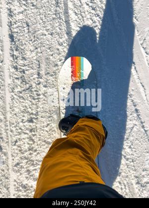 Point-of-View-Aufnahme eines Snowboarders, der gelbe Hosen und schwarze Stiefel auf einem farbenfrohen Snowboard trägt, mit einem langen Schatten über eine sonnendurchflutete schneebedeckte Piste Stockfoto