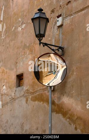 Eine Straßenlaterne aus schwarzem Metall und ein konvexer Verkehrsspiegel sind an einer stark strukturierten, hellbraunen Wand mit abgebrochener Farbe montiert. Der Spiegel reflektiert Stockfoto