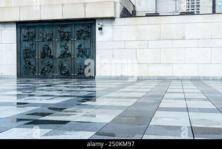 Eine Nahansicht einer strukturierten Steinmauer mit einer großen Bronzetür mit komplizierten Reliefskulpturen. Der Boden ist aus polierten Fliesen reflektiert Stockfoto