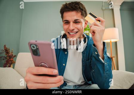Junger Mann lächelt beim Online-Shopping mit Smartphone und Kreditkarte, um auf einem Marktplatz zu kaufen, in einem gemütlichen Wohnzimmer. Glücklicher Kerl, der eine Lieferung mit einer Handy-App bestellt. Hochwertige Fotos Stockfoto