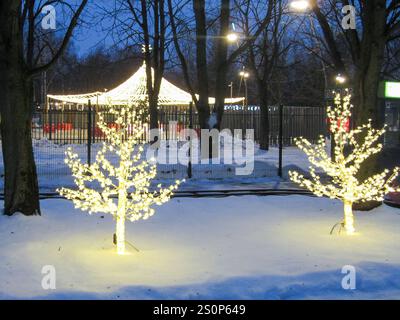 Zwei beleuchtete Bäume leuchten in einem verschneiten Park mit einem Karussell im Hintergrund, umgeben von einem Zaun und Abendlichtern, die an eine gemütliche Winterszene erinnern Stockfoto