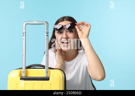 Frau mit Koffer, die Sonnenbrille auf hellblauem Hintergrund trägt Stockfoto