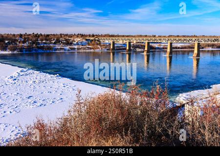 Eine Brücke überspannt einen Fluss mit einem schneebedeckten Ufer auf beiden Seiten. Die Brücke ist von Bäumen und Büschen umgeben. Das Wasser ist ruhig und klar Stockfoto