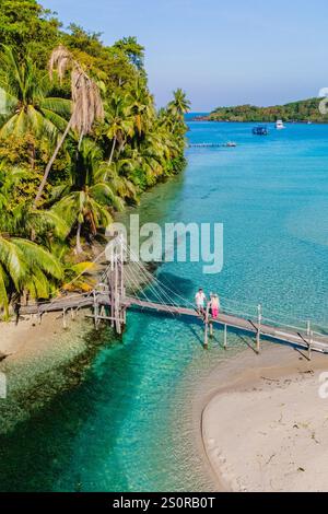 Üppige Palmen umgeben eine ruhige Küstenlinie, während zwei Reisende entlang einer Holzbrücke spazieren und die atemberaubende Aussicht auf Koh Koods kristallklares Wasser und die lebhafte Umgebung genießen. Stockfoto