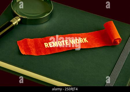 Auf einem dunklen Notebook befindet sich ein leuchtend orangefarbenes Band mit der Aufschrift Remote Work. In der Nähe befindet sich eine Lupe, die auf Fokus und Organisation hindeutet Stockfoto
