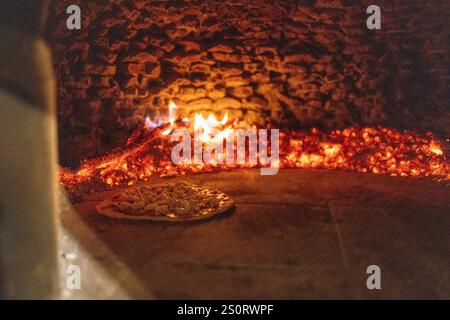Eine Pizza voller Beläge, die in einem traditionellen Holzofen gebacken wird, umgeben von glühender Glut und Flammen. Stockfoto