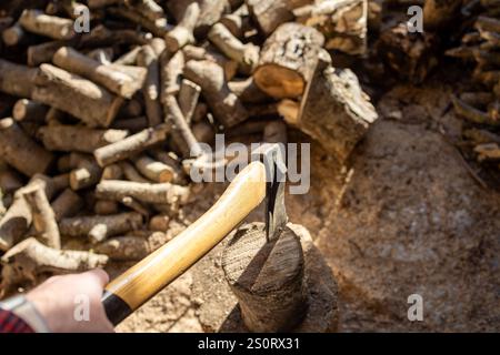 Eine Nahaufnahme einer Person, die eine Axt hält, während sie einen Baumstamm auf einem Baumstumpf spaltet, umgeben von einem Haufen Brennholz und Sägemehl. Stockfoto