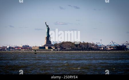 Ein Fernblick auf die Freiheitsstatue auf Liberty Island, umgeben von Wasser. Der Himmel ist klar mit ein paar Wolken, und die Skyline der Stadt ist f Stockfoto