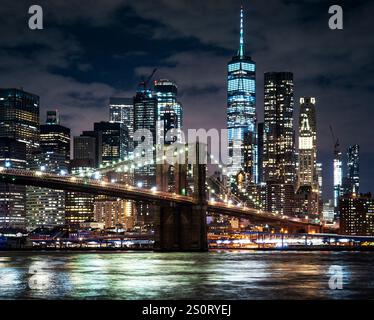 Ein atemberaubender Blick auf die Brooklyn Bridge bei Nacht mit der Skyline von Manhattan im Hintergrund, mit beleuchteten Wolkenkratzern und einem reflektierenden Fluss. Stockfoto