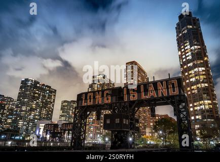 Ein nächtlicher Blick auf Long Island City mit modernen Wolkenkratzern, die vor einem bewölkten Himmel beleuchtet werden. Das legendäre Long Island-Schild steht im Th Stockfoto