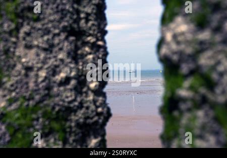 Ein ruhiger Blick auf ein Segelboot auf dem Wasser, eingerahmt von zwei felsigen Strukturen, die mit Moos bedeckt sind. Die Szene fängt die Ruhe des Strandes und des dist ein Stockfoto