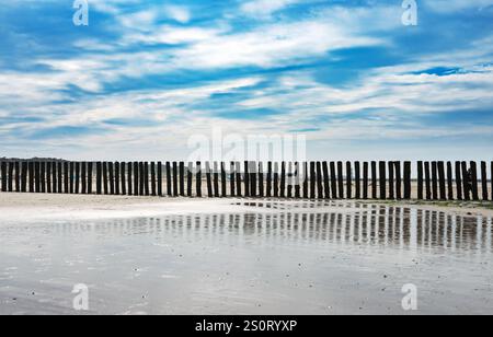 Eine ruhige Strandszene mit Holzpfosten, die in einer Reihe an der Küste stehen und sich im nassen Sand spiegeln. Der Himmel ist teilweise bewölkt mit einem blauen hu Stockfoto