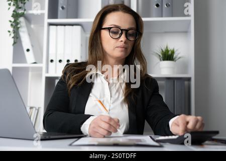 Eine engagierte, professionelle Frau arbeitet an einem Schreibtisch und nutzt ihren Laptop und Taschenrechner, um verschiedene Aufgaben effektiv zu erledigen und zu präsentieren Stockfoto