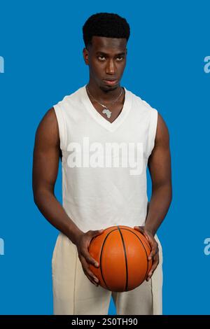 Studio-Porträt eines ernsthaften Basketballspielers, der einen Ball hält, ein weißes Hemd und einen afrikanischen Anhänger trägt, auf einem leuchtend blauen Hintergrund Stockfoto