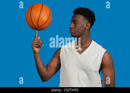 Junger Mann, der einen Basketball auf der Fingerspitze vor einem leuchtend blauen Hintergrund dreht Stockfoto
