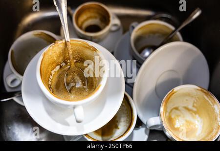 Gebrauchte Kaffeetassen und Löffel, die nach dem Kaffeekosum in einem Spülbecken gestapelt wurden Stockfoto