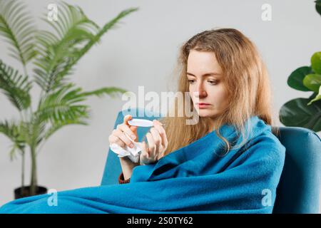 Eine junge Frau, in eine blaue Decke gewickelt, sitzt auf einer Couch und sieht ein Thermometer mit besorgtem Ausdruck an. Sie scheint sich unwohl zu fühlen, umgeben von Stockfoto