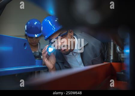Maschinenbauingenieur, afroamerikanischer Mann, der Taschenlampe für die Maschinensteuerung benutzt, arbeitet in einem Team mit einer indischen Kollegin. Stockfoto