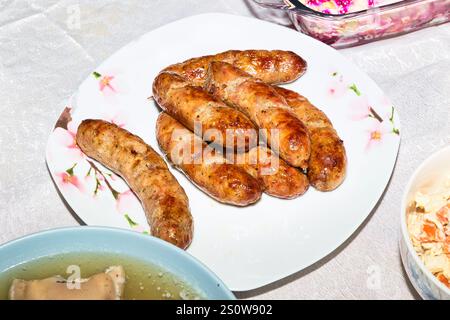 Eine weiße quadratische Platte bietet Platz für mehrere gegrillte Würstchen. Andere Gerichte, darunter eine Schüssel Salat und ein Gericht mit Brühe, sind auf einem hellen Tisch zu sehen Stockfoto