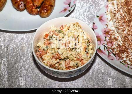 Auf einem Tisch steht eine Schüssel mit russischem Salat, Tellern mit gegrillten Würstchen und einem Dessert mit Schokoladenspänen. Die Einstellung scheint ein Casu zu sein Stockfoto