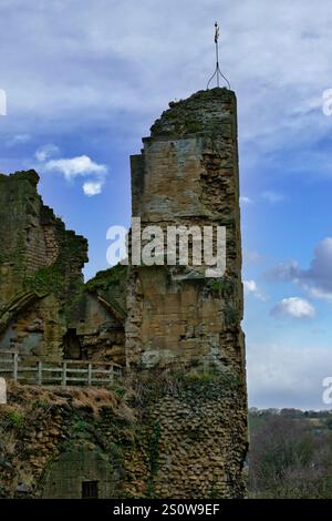 Teilweise ruinierter Steinturm mit starker Verwitterung und Vegetation. Ein Metallfinial übertrifft die Struktur vor einem teilweise bewölkten blauen SK Stockfoto