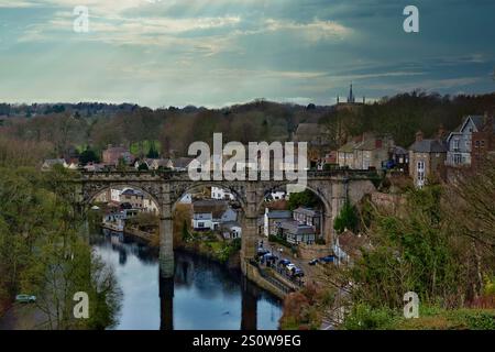 Das Steinviadukt überspannt einen Fluss und überblickt ein Dorf mit Häusern, einem Kirchturm und bewaldeten Hügeln unter bewölktem Himmel. Die Szene ist ruhig und eigenartig Stockfoto