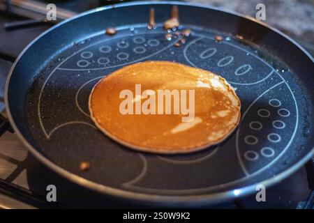 Ein einzelner, goldbrauner Pfannkuchen kocht in einer dunklen Antihaftpfanne auf einem Herd. Die Pfanne hat ein dekoratives Muster. Ölrückstände sind sichtbar. Stockfoto