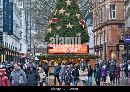 Eine geschäftige Straßenszene in der Stadt an einem regnerischen Tag zeigt einen großen Weihnachtsbaum mit Sternen und Herzen, einen Marktstand mit einer digitalen Werbung Stockfoto