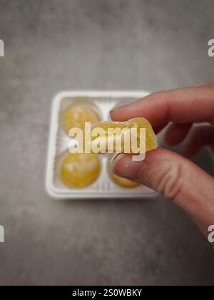 Hand hält ein halb gegessen Stück Mango Mochi Dessert über einem offenen Paket auf grauem Hintergrund in prag, tschechien Stockfoto