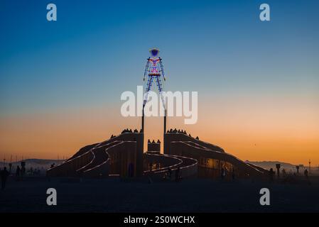 Eine große, farbenfrohe humanoide Skulptur steht auf einer Holzkonstruktion in der Black Rock Desert bei Sonnenuntergang, umgeben von Festivalteilnehmern. Stockfoto