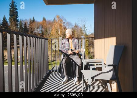 Der junge Musiker sitzt auf einem sonnigen Balkon, in der Ukulele. Umgeben von Herbstbäumen genießt er den friedlichen Moment im Freien, verloren in h Stockfoto