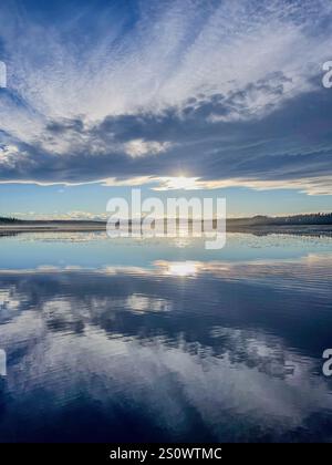 Ein stiller See, der einen lebendigen Himmel mit Sonnenlicht reflektiert, das durch Wolken schaut. Perfekt für friedliche und stimmungsvolle Landschaften. Stockfoto
