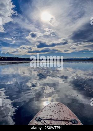 Ein Paddleboard schwingt auf einem ruhigen See und spiegelt dramatische Wolkenformationen und Sonnenlicht unter einem blauen Himmel wider. Ideal für Natur, Abenteuer und Outdoor-Leben Stockfoto