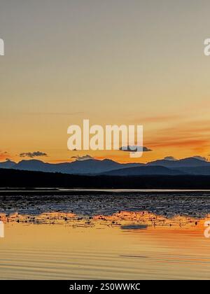 Ein ruhiger See spiegelt die lebendigen Farben eines Sonnenuntergangs wider, umgeben von fernen Berggipfeln und einer ruhigen Wasserlandschaft. Stockfoto