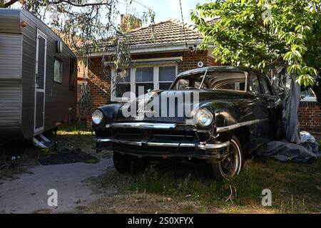 Alte schwarze Chrysler Windsor Limousine, die in einem schattigen Vorgarten sitzt, in einem Zustand der Verwüstung, neben einem Wohnwagen, mit einem Vorstadthaus im Hintergrund Stockfoto