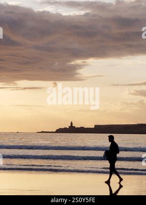 Surfer in Silhouette, der mit seinem Brett am Ufer mit Insel dahinter bei Sonnenuntergang in der Stadt Essaouira spaziert, 29. Dezember 2024 Stockfoto