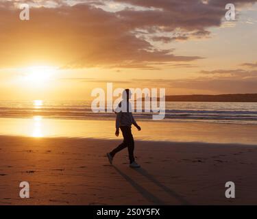 Schöner junger Mann mit Sonnenbrille und langen Haaren spaziert entlang des Sandstrandes mit Meer und Insel hinter dem Sonnenuntergang in Essaouira, 29. Dezember 2024 Stockfoto