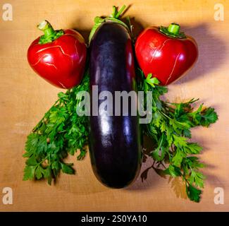 Farbenfrohe rote Paprika und eine glänzende Aubergine werden künstlerisch mit frischem Koriander auf einer Holzoberfläche platziert. Dieses kreative Arrangement hebt die Be hervor Stockfoto