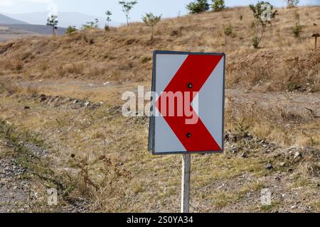 Das hellrot-weiße Straßenschild warnt Fahrer vor einer bevorstehenden scharfen Linkskurve in einer trockenen, kargen Landschaft unter klarem Himmel. Stockfoto