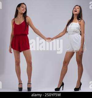 Zwei wunderschöne Zwillingsschwestern, gekleidet in stilvollen Playsuits und High Heels, halten die Hände, während sie mit neutralen Ausdrücken in die Kamera blicken, die El ausstrahlen Stockfoto