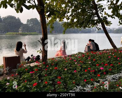 Eine Frau in traditioneller Kleidung fotografiert den Sonnenuntergang am Hoan Kiem Lake und fängt das tägliche Leben in Hanoi, Vietnam, ein. Stockfoto