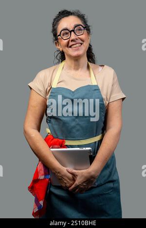 Glückliche Küchenchefin mit Schürze, Tablette und Blick nach oben, zeigt Selbstvertrauen und Positivität in kulinarischen Fähigkeiten Stockfoto