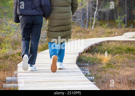 Ein Paar, das auf einem Holzsteg im Wald spaziert. Die Frau trägt eine grüne Jacke, und der Mann trägt eine schwarze. Beide tragen Sneake Stockfoto