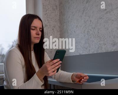 Junge Frau, die sich auf den Smartphone-Bildschirm konzentriert, Kreditkarte greift, Online-Banking-Transaktionen in gemütlicher häuslicher Umgebung abschließt Stockfoto