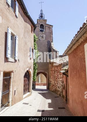 Enge Gasse mit farbenfrohen Gebäuden, die an einem sonnigen Tag zu einem historischen Glockenturm in roussillon, frankreich, führt Stockfoto